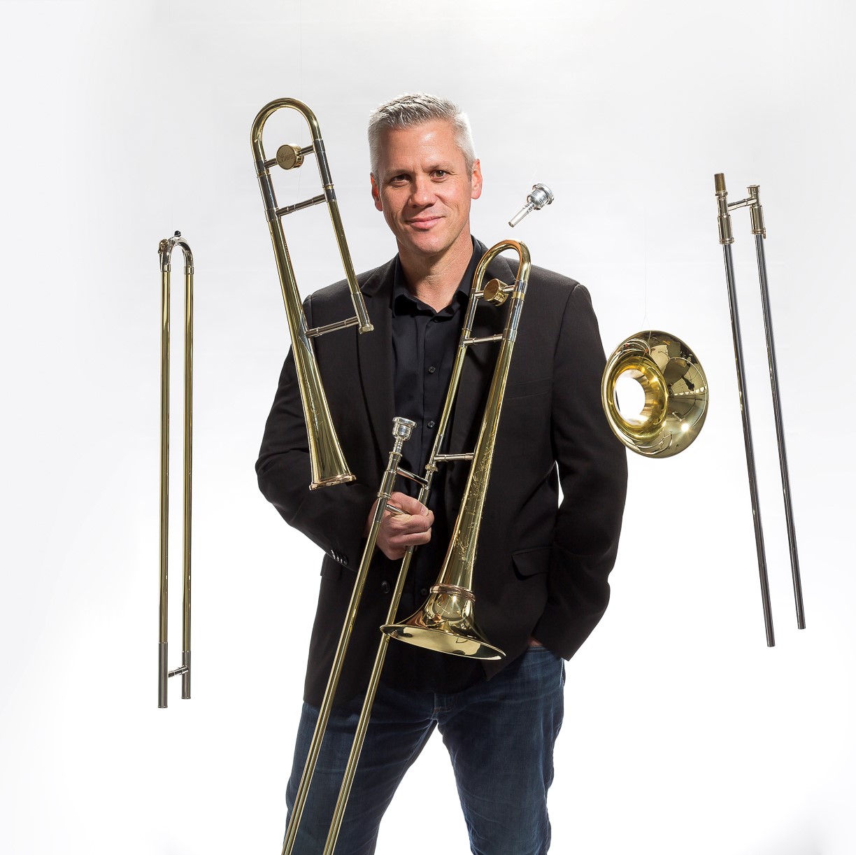 A smiling man stands holding a trombone as trombone parts float around him