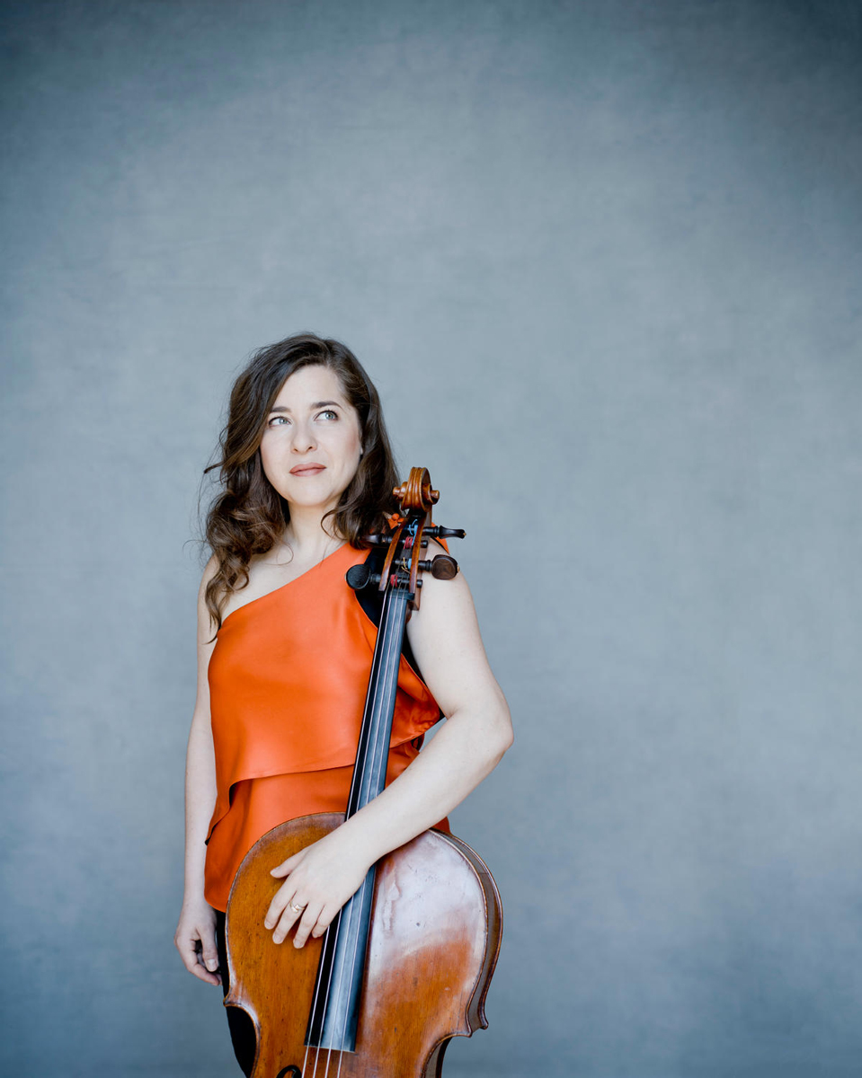 A woman with long brown hair wearing an orange dress stands with a cello