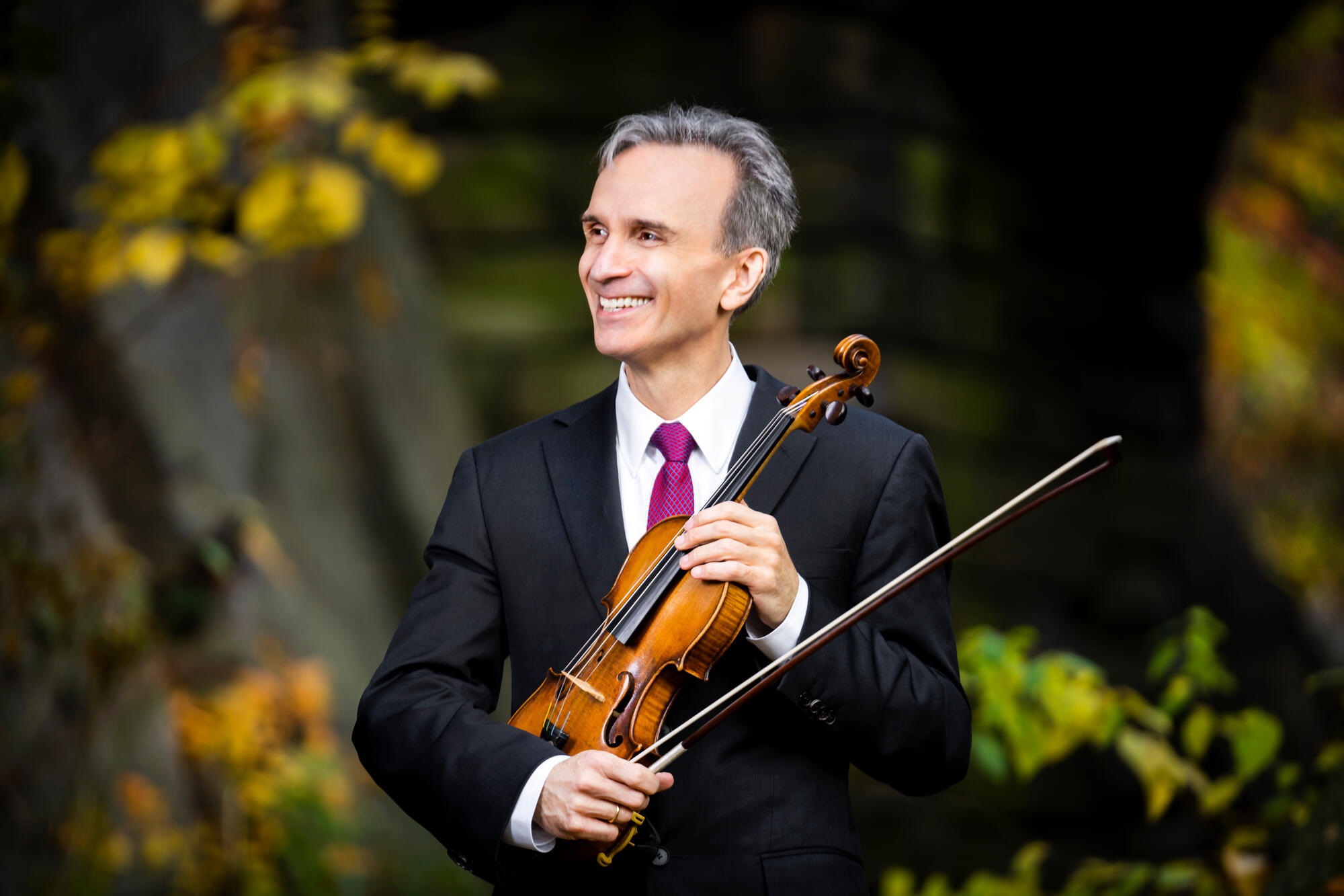 Gil Shaham with violin smiling in a suit and tie, trees behind